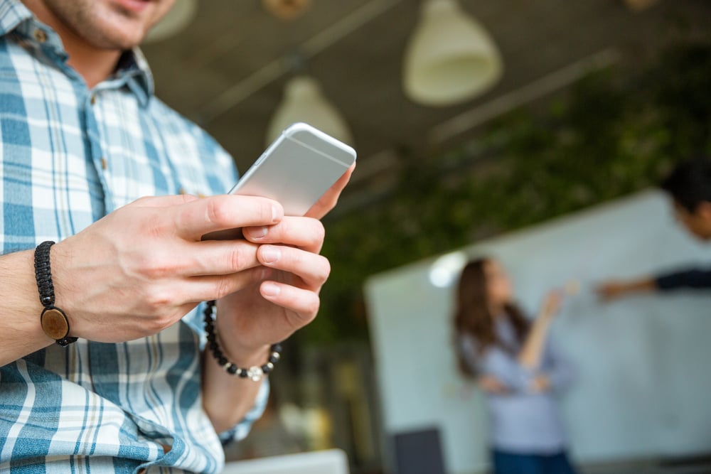 Closeup of hands of young man in checkered shirt using mobile phone while his partners arguing-2