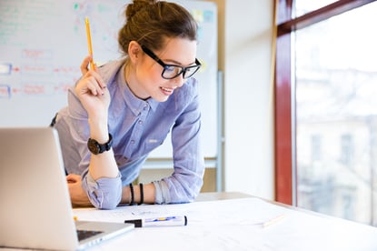 Happy young woman in glasses standing near the window in office and working with blueprint-1