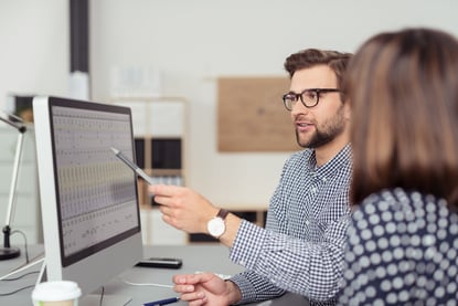 Proficient young male employee with eyeglasses and checkered shirt, explaining a business analysis displayed on the monitor of a desktop PC to his female colleague, in the interior of a modern office-4